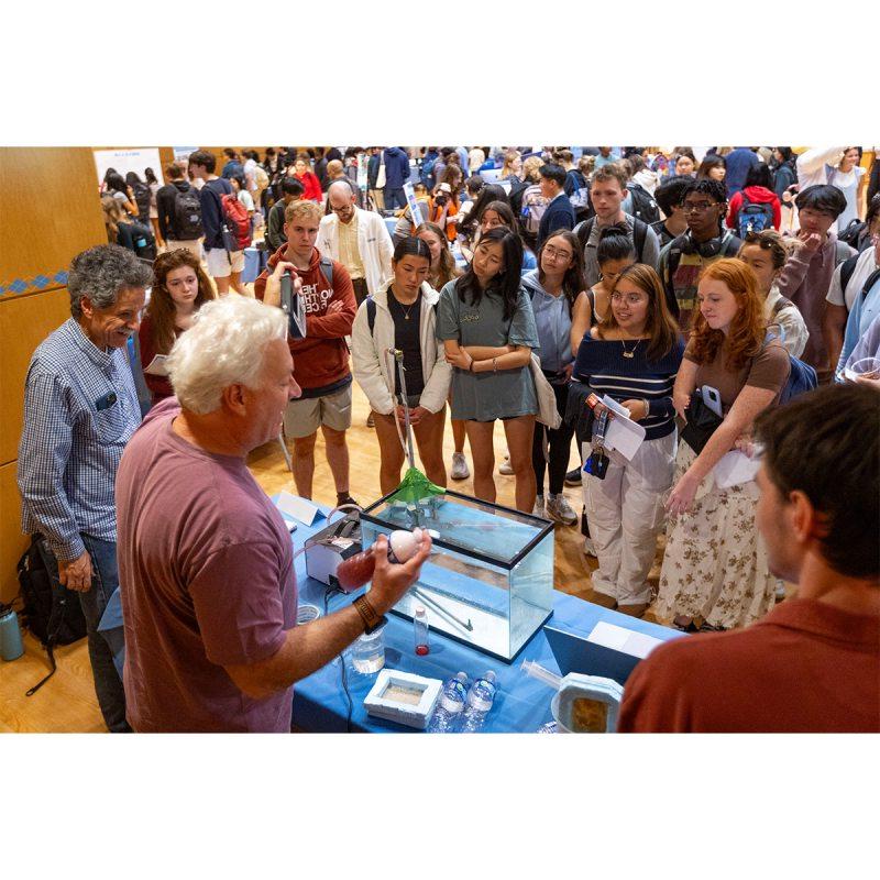 Professors talking to a group of students at a Research Week fair on the campus of UNC-Chapel Hill inside of a hall.